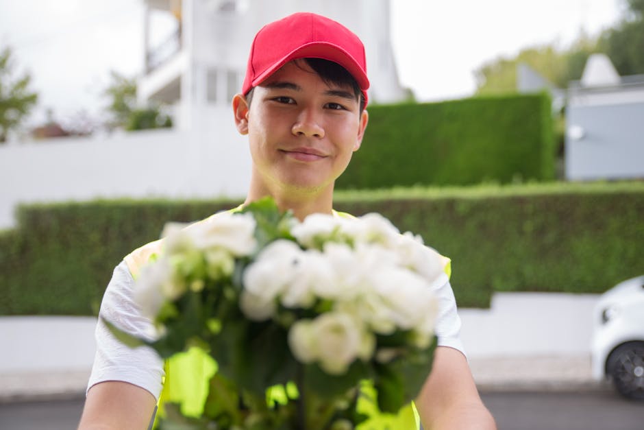 A delivery person, wearing a red cap and yellow high-visibility vest, presents a bouquet of fresh white roses and lilies, with green foliage, wrapped in clear cellophane, for a floral gift or flower d