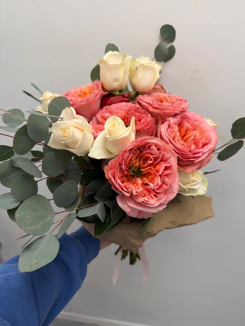 A hand in a blue sleeve holds a bouquet of fresh flowers wrapped in brown paper, featuring large pink ranunculus with ruffled petals, creamy white roses with tightly closed buds, and eucalyptus leaves